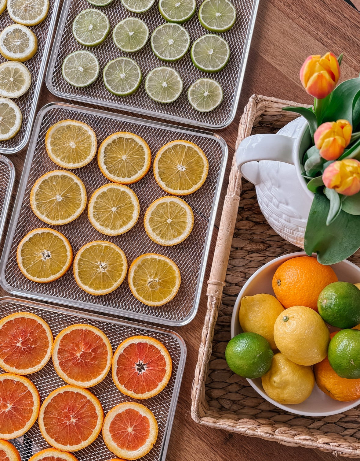 Trays of dehydrated citrus fruits on a wooden table with a bowl of fresh fruits and flowers.